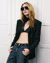 Mannequin portant un blazer noir à laçage, un soutien-gorge noir ajouré, un jean taille haute, des lunettes de soleil oversize et un foulard en cuir noué autour du cou, sur un fond à panneaux blancs. | Pinko