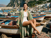 Una donna sorridente in kimono tie-dye verde, top a costine bianco e shorts, con sandali Pinko a stampa pitone, seduta su una barca da pesca nel porto di Capri. | Pinko