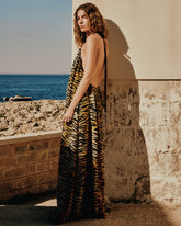 Una donna in lungo abito animalier con schiena scoperta posa contro un muro in pietra illuminato dal sole, con una costa rocciosa e il mare blu sullo sfondo. | Pinko