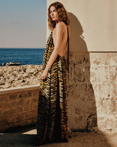 Una donna in lungo abito animalier con schiena scoperta posa contro un muro in pietra illuminato dal sole, con una costa rocciosa e il mare blu sullo sfondo. | Pinko