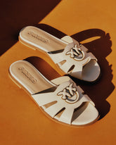 Un paio di sandali flat Pinko in pelle bianca con fascetta traforata e logo rondine in metallo dorato, su superficie arancione. | Pinko