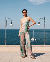 Una donna in coordinato di raso tie-dye verde salvia e rosa composto da canotta e pantaloni a gamba larga, occhiali da sole in tartarugato e sandali flat Pinko color cuoio, in piedi su un lungomare con una barca a vela sullo sfondo. | Pinko